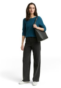 Schwarze Lederhandtasche, türkisfarbener gerippter Pullover, schwarze weit geschnittene Jeans, weiße Sneaker. Model steht vor einem schlichten weißen Hintergrund.