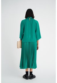 Abito plissettato verde con collo alto e schiena con apertura a chiave, abbinato a calze nere e sandali, mentre tiene una pochette beige.