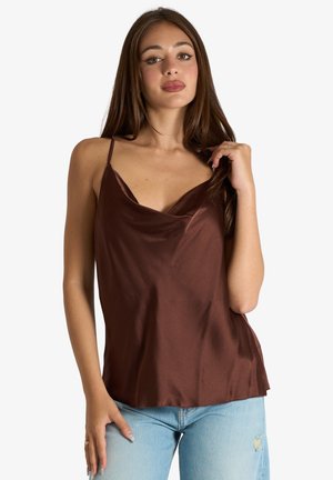 Giovane donna con lunghi capelli castani che indossa un top senza maniche in raso marrone e jeans azzurri strappati su sfondo bianco.
