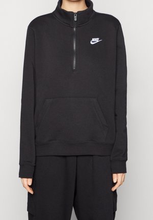 Zwarte Nike half-zip pullover met voorzak, gedragen door een persoon in bijpassende zwarte broek tegen een effen achtergrond.