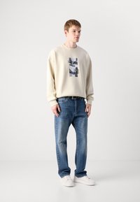 Beige genser med grafisk trykk, paret med blå denimbukser. Modellen bruker hvite sneakers. Uformell passform med avslappede silhuetter.