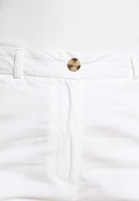 Pantalones de algodón blancos con una textura suave, que presentan un cierre de botón de cuerno y costuras de hilo en contraste a lo largo de la cintura y los bolsillos.
