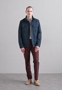 Jachetă închisa la culoare, cu două buzunare pe piept, peste o cămașă cu model floral, asortată cu pantaloni burgundii conici și pantofi bej. Textură netedă.