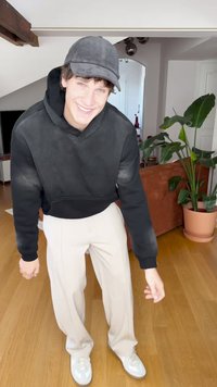 Black hooded sweatshirt, beige wide-leg trousers, and light sneakers. Indoor setting with a potted plant and brown couch in background.