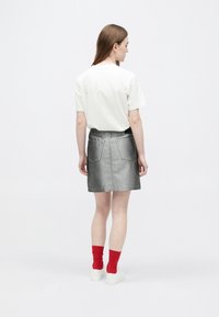 Fehér pólót, szürke farmer szoknyát, piros zoknit és fehér tornacipőt viselő nő áll háttal egy egyszínű háttér előtt.