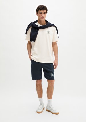 Jeune homme debout portant un t-shirt blanc, un short bleu marine avec le numéro 67, des baskets blanches et un sweat bleu marine posé sur les épaules.
