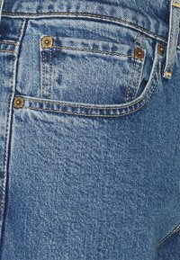 Zblízka pohled na modré džíny ukazující zadní kapsu s viditelnými měděnými nýtky a detailním prošitím.