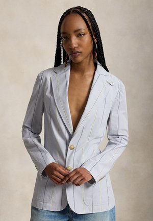 Žena s dlhými copy nosiaca svetlosivý pruhovaný blazer s béžovými gombíkmi, stojaca pred neutrálnym pozadím.