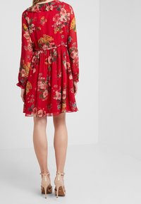 Robe rouge à fleurs avec un col à volants et des manches longues, dotée d'une taille froncée. Associée à des chaussures à talons hauts nude.