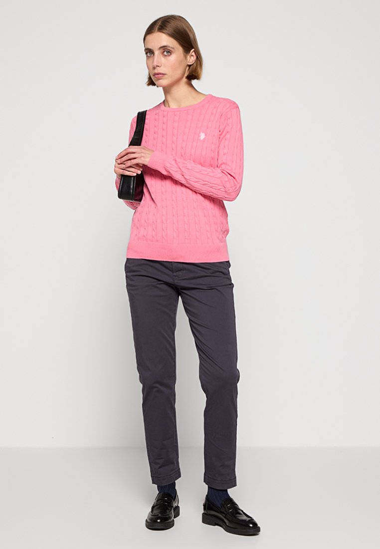 Rosa Kabelstrickpullover mit Rundhalsausschnitt, kombiniert mit dunkelgrauen Hosen und schwarzen Schuhen. Das Model hält eine schwarze Handtasche.