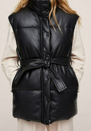 Veste sans manches en cuir noir matelassé avec boutons-pression à l'avant et ceinture nouée, portée sur un haut léger à manches longues, mains dans les poches.