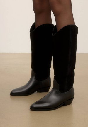 Bottes noires montant jusqu'au genou avec petits talons portées sur jambes nues, montrées sur un sol beige uni et un fond clair.
