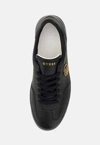 Sneaker in pelle nera con punta rotonda, superficie strutturata, lacci piatti neri, logo dorato "GUESS" e emblema decorativo rotondo sul lato.