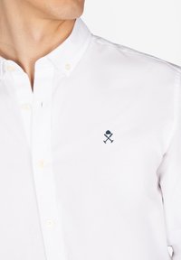 Camisa blanca de botones hecha de algodón con una textura suave. Presenta un logo bordado en color navy en el lado izquierdo del pecho y un diseño de cuello estándar.