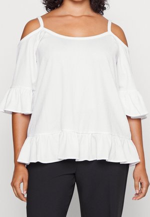 Femme portant un haut blanc épaules dénudées avec des manches et un ourlet à volants, associé à un pantalon noir, présenté du cou aux hanches.