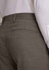 Brązowe spodnie w kratę z jednolitym paskiem, wyposażone w tylną kieszeń i wykonane z teksturowanego materiału. Połączone z białą koszulą.