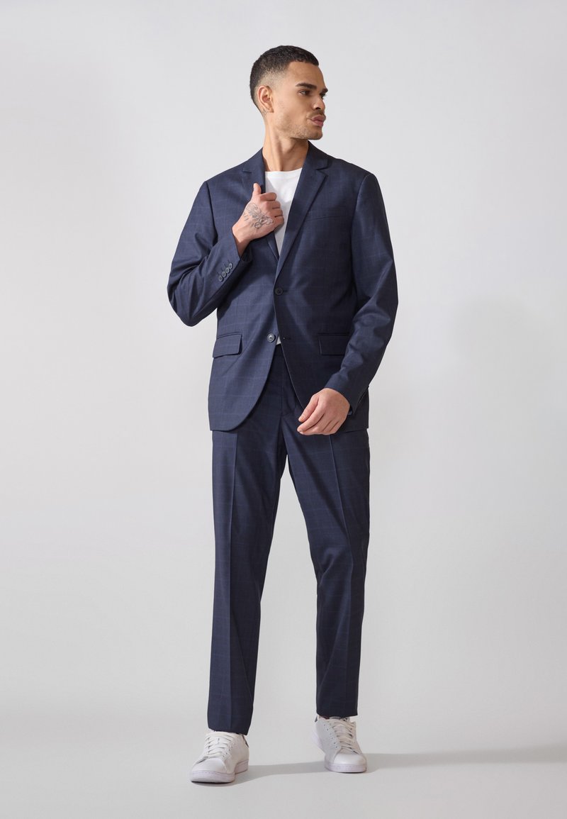 Costume à carreaux bleu marine avec une veste sur mesure et un pantalon assorti, associé à une chemise blanche et des baskets blanches, présentant un design élégant.