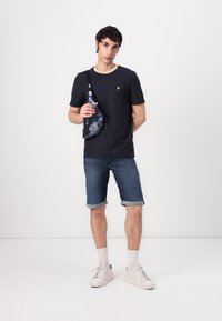Tricou bleumarin cu guler rotund și logo galben, pantaloni scurți din denim, adidași albi și o geantă de umăr cu model albastru.