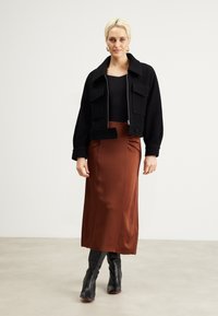 Svart cropped jakke med store lommer, kombinert med et langt, rustfarget satengskjørt og knelang svarte støvler på en nøytral bakgrunn.