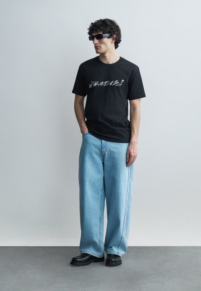 Άνδρας με κατσαρά μαλλιά που φοράει σκούρα γυαλιά ηλίου, μαύρο κοντομάνικο με στάμπα, φαρδιά ανοιχτό μπλε τζιν και μαύρα παπούτσια, στέκεται μπροστά σε έναν γκρι τοίχο.