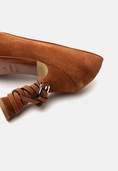 Καφέ δερμάτινο γυναικείο παπούτσι με τακούνι, μυτερό μύτη και λουράκι που τυλίγεται γύρω από τον αστράγαλο με ασημένιες αγκράφες.