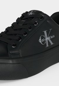 Черен нисък кец с черни връзки, гумена подметка и лого Calvin Klein, отпечатано в бяло отстрани и на езика.