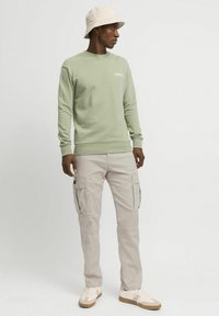 Lysegrønn genser med ribbestrikkede mansjetter, beige cargobukser med sidelommer, og en beige bøttehatt. Hvite joggesko fullfører antrekket.