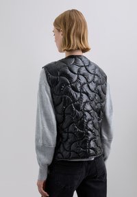 Black quilted vest featuring shiny texture and scalloped pattern, worn over a gray long-sleeve shirt, with a round neckline and casual fit.