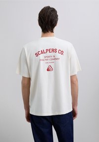 Camiseta de algodón color crema con mangas cortas, con texto rojo "SCALPERS CO. SPORTY & HEALTHY COMPANY COOL PLAYERS" en la parte trasera.