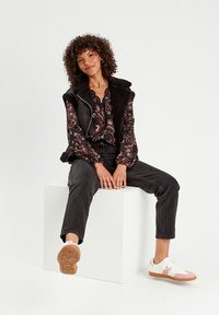 Gilet foderato in shearling nero sopra una blusa stampata, abbinato a jeans neri a vita alta e sneakers bianche con dettagli rosa.