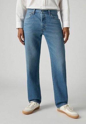 Jeans rectos de color azul claro con una sutil textura vertical, que cuentan con cinco bolsillos y cierre de botón, combinados con zapatillas blancas.