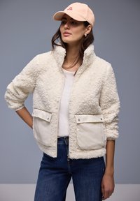 Femme portant une veste sherpa de couleur crème avec de grandes poches avant, une chemise blanche, un jean bleu et une casquette de baseball beige.