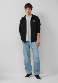 Zwarte ritsjack over een grijs t-shirt, gecombineerd met lichtblauwe versleten jeans en zwarte sneakers. Eenvoudig ontwerp met een casual pasvorm.