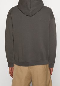 Una persona che indossa una felpa con cappuccio grigia, vista di spalle, con pantaloni beige e le mani ai lati contro uno sfondo neutro.