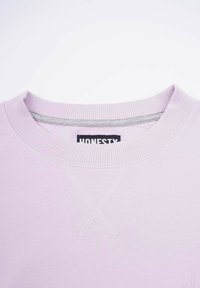 Ljuslila sweatshirt med ribbat runt halsen och förstärkande sömmar i ett X-mönster. Etiketten säger "ÄRLIGHET". Mjuk tygstruktur.