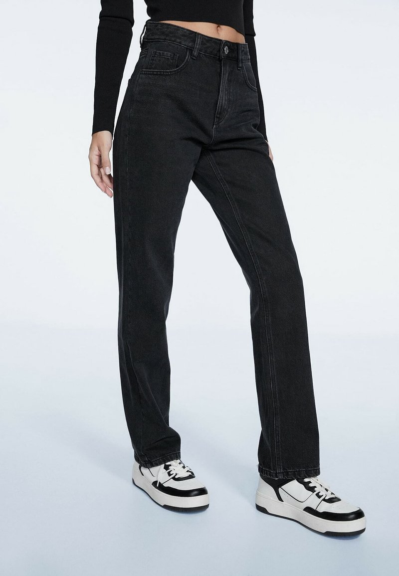 Personne portant un jean noir taille haute à jambes droites et des baskets noir et blanc, debout devant un fond clair uni.