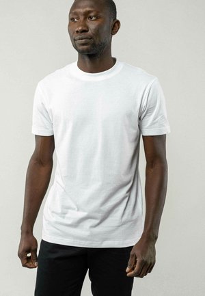 Weißes Baumwoll-T-Shirt mit kurzen Ärmeln und rundem Ausschnitt, getragen von einer Person in schwarzen Hosen. Glatte Textur, klassischer Schnitt.