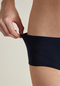Tmavě modré bavlněné slipy, které jsou taženy za páskem, což zdůrazňuje jejich měkkou texturu, elastický materiál a přiléhavý design. Kůže viditelná pod páskem.