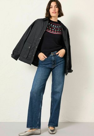 Dunkelblaue Jacke mit Knöpfen, schwarzer Pullover mit rotem und weißem Muster im Schulterbereich, kombiniert mit hellblauen weiten Jeans und silbernen Ballerinas.