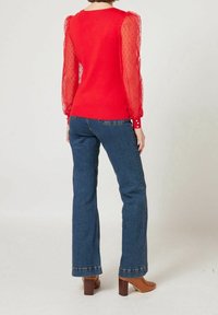 Pull rouge avec manches en tulle à pois, associé à un jean flare bleu et des bottines marron à talons blocs. Texture douce, coupe ajustée.