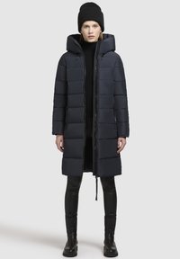 Navy blue puffer coat with a hood, featuring a zip front, quilted design, and side pockets, paired with black leggings and a knit beanie.