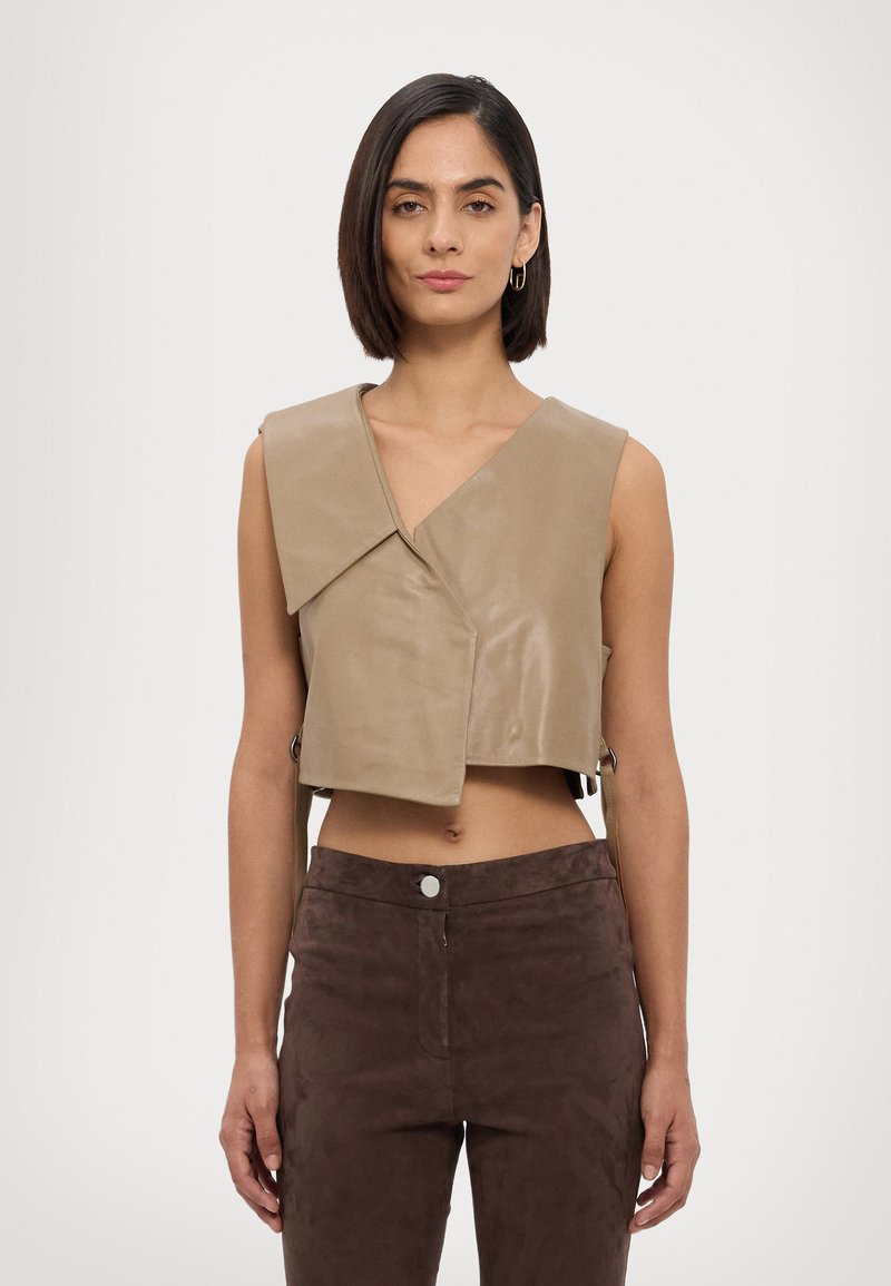 Beige, ärmelloses, kurzes Top aus glattem Leder mit V-Ausschnitt und asymmetrischem Vorderdesign. Kombiniert mit braunen Wildleder-Hosen mit hoher Taille.