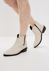 Botas de tobillo de cuero blanco con costuras decorativas, puntera puntiaguda, cremallera lateral y un pequeño tacón apilado. Apariencia texturizada.