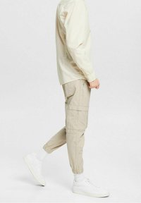 Persoon in beige cargopantalon, witte sokken, witte sneakers en een lange beige shirt, die met één voet opgetild in de middenstap loopt.