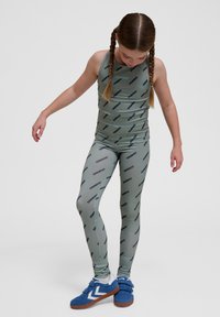 Grön träningsoverall bestående av en ärmlös topp och matchande leggings, båda med ett upprepande mönster av varumärkeslogotypen; bärs med blå sneakers.