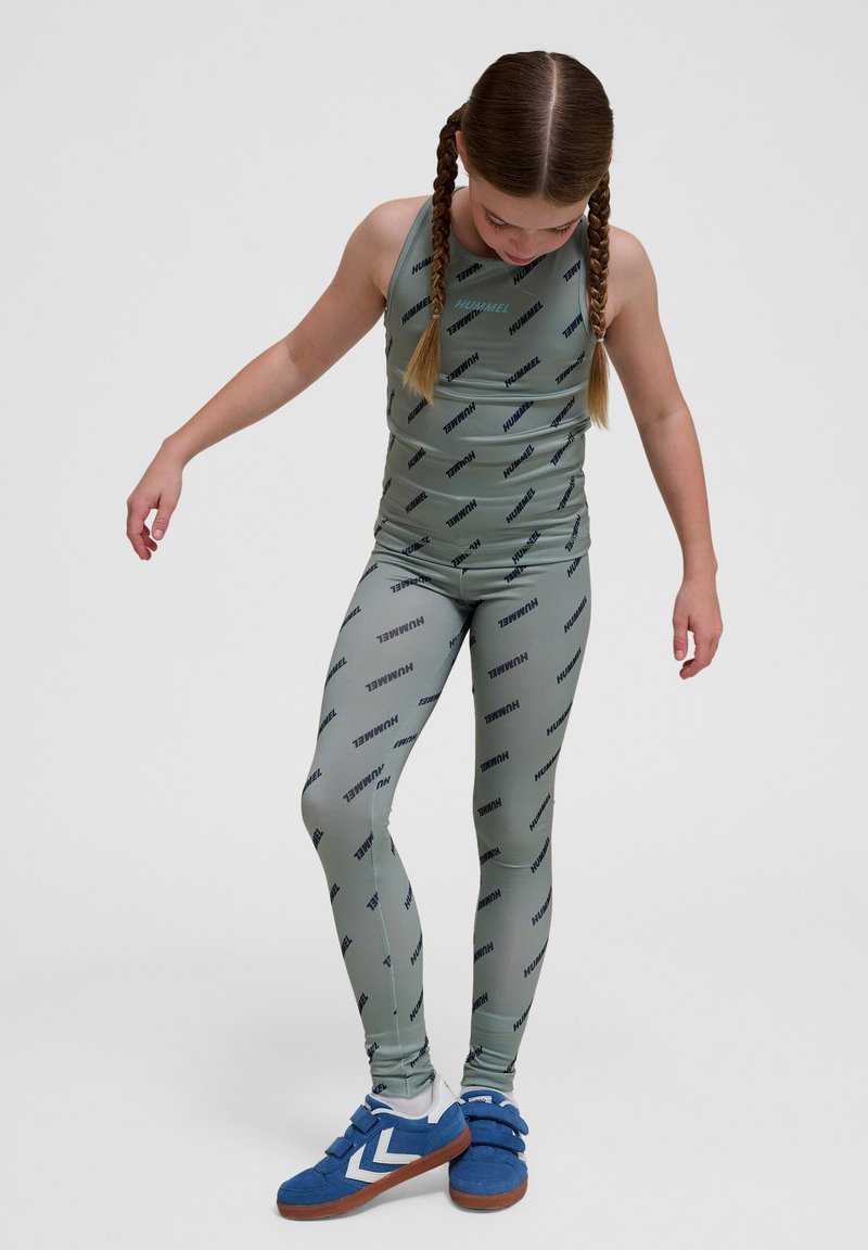 Grön träningsoverall bestående av en ärmlös topp och matchande leggings, båda med ett upprepande mönster av varumärkeslogotypen; bärs med blå sneakers.