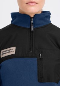 Giacca in pile nera e blu con colletto alto, zip frontale e tasca laterale. Presenta un'etichetta di stoffa sul petto.