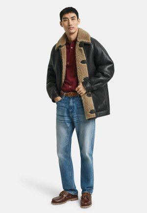 Homme portant une veste en shearling noire sur un polo rouge, un jean bleu, une ceinture marron et des chaussures en cuir marron, se tenant les mains dans les poches.
