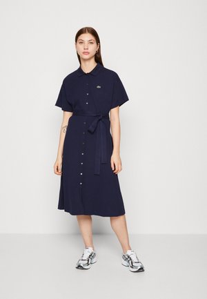 Vestido de manga corta azul marino con cierre de botones en la parte delantera y un lazo en la cintura. Presenta un pequeño logo verde y una falda acampanada. Combinado con zapatillas plateadas.