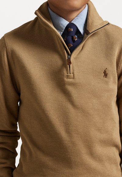 Καφέ πουλόβερ με κουμπί quarter-zip μπροστά, ribbed υφή και κεντημένο λογότυπο. Φοριέται πάνω από ένα ανοιχτό μπλε πουκάμισο με γιακά και σχέδιο γραβάτα.